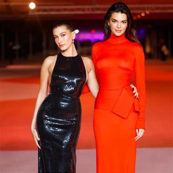 Hailey Bieber and Kendall Jenner pose on a sandy beach in matching cream and white gowns
